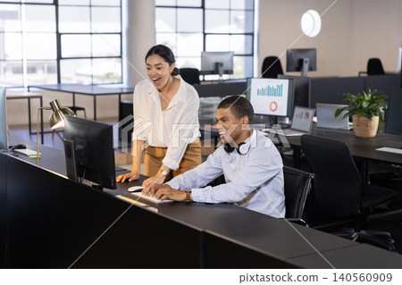 Diverse coworkers collaborating on report at open-plan office desk, monitor, lamp, headphones 140560909