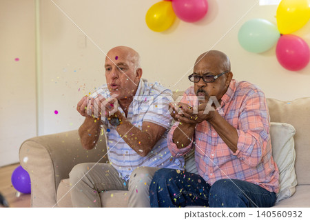 Diverse senior male duo blowing colorful confetti on beige sofa at home with pastel balloons Diverse senior male duo blowing colorful confetti on beige sofa at home with pastel balloons 140560932