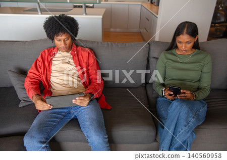 Diverse couple sitting on grey sofa in living room scrolling smartphone, browsing tablet Diverse couple sitting on grey sofa in living room scrolling smartphone, browsing tablet 140560958
