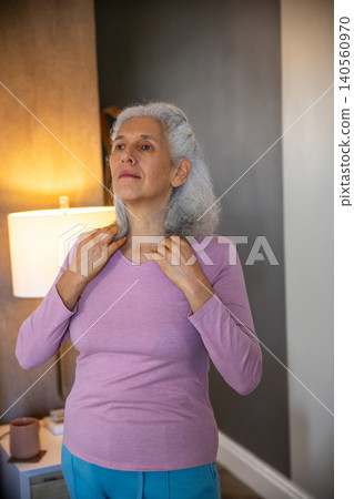 Senior woman adjusting lavender shirt while standing at home beside side table with lamp and mug Senior woman adjusting lavender shirt while standing at home beside side table with lamp and mug 140560970