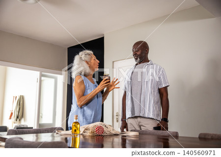 Diverse senior couple holding jar of preserve and standing by wooden dining table with mesh bag 140560971