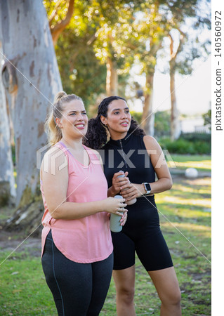 Female friends standing holding reusable water bottles in sunlit city park with smartwatch Female friends standing holding reusable water bottles in sunlit city park with smartwatch 140560972