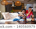 Diverse male friends sitting on sofa holding controllers while sharing popcorn in living room 140560979