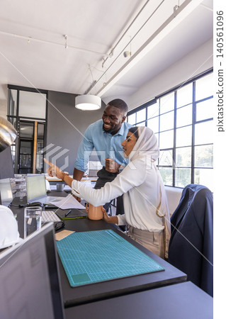 Diverse coworkers pointing at laptop screen, holding coffee with cutting mat at desk in office Diverse coworkers pointing at laptop screen, holding coffee with cutting mat at desk in office 140561096