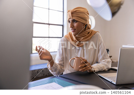 Mid adult indian woman holding mug, pointing at plans beside silver laptop at studio desk Mid adult indian woman holding mug, pointing at plans beside silver laptop at studio desk 140561104