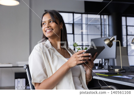 Asian woman sitting at desk in modern office holding tablet and smiling near computer monitors Asian woman sitting at desk in modern office holding tablet and smiling near computer monitors 140561108