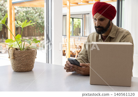 Mid adult indian man in turban sitting at kitchen island checking smartphone near plant, sealed box Mid adult indian man in turban sitting at kitchen island checking smartphone near plant, sealed box 140561120