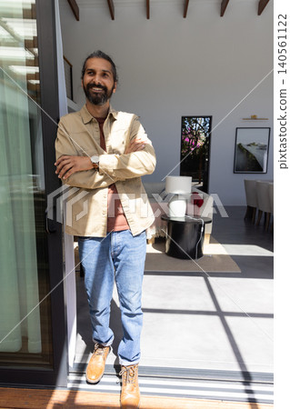 Mid adult indian man standing in sliding glass doorway, leaning on door frame wearing wristwatch 140561122