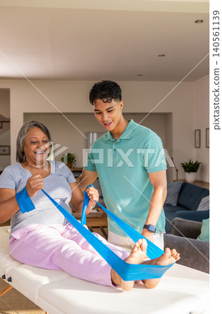 Senior african american patient, hispanic therapist working with band on table at home Senior african american patient, hispanic therapist working with band on table at home 140561139