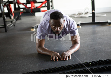 Mid-adult African American man in sportswear holding plank position on gym floor with battle rope Mid-adult African American man in sportswear holding plank position on gym floor with battle rope 140561163