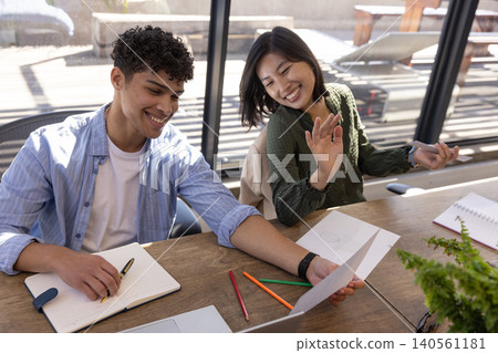 Diverse coworkers reviewing printed chart in office at table with laptop, notebooks and potted fern Diverse coworkers reviewing printed chart in office at table with laptop, notebooks and potted fern 140561181