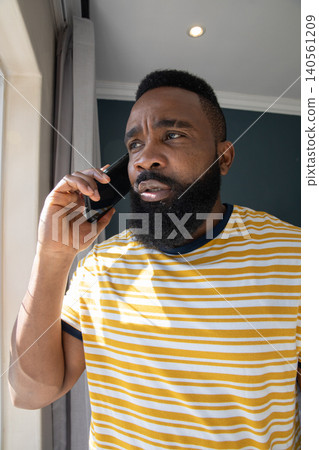 African American man talking on smartphone standing by window with gray curtain and striped t-shirt 140561209