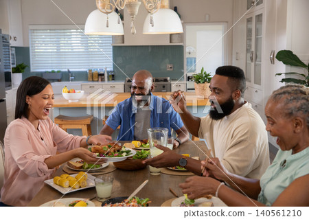 Diverse extended family passing dishes and serving salad at kitchen dining table with roast chicken Diverse extended family passing dishes and serving salad at kitchen dining table with roast chicken 140561210