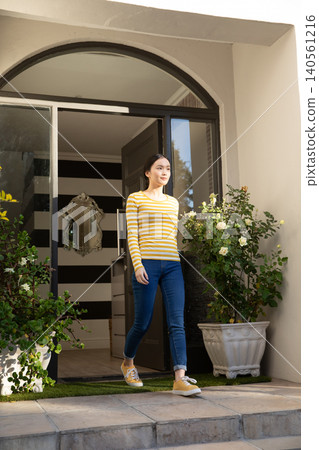 Asian woman stepping through front door onto porch past turf, planters, striped wall mirror Asian woman stepping through front door onto porch past turf, planters, striped wall mirror 140561216
