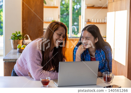 Asian mother and daughter sitting in modern kitchen at island using silver laptop while sipping tea 140561266