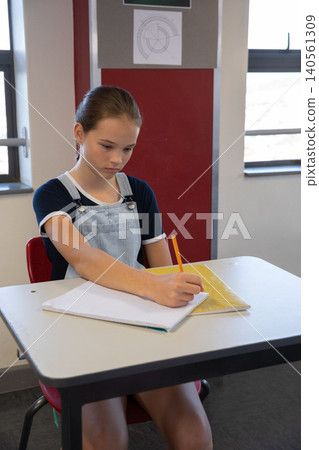 Female teenager writing on notebook over folder at desk in classroom with geometry poster Female teenager writing on notebook over folder at desk in classroom with geometry poster 140561309