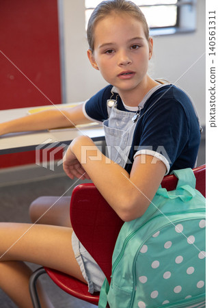 Girl student sitting at school desk holding red chair while backpack hanging from chair Girl student sitting at school desk holding red chair while backpack hanging from chair 140561311