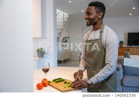 African american man in apron slicing zucchini on cutting board at kitchen island, copy space African american man in apron slicing zucchini on cutting board at kitchen island, copy space 140561330