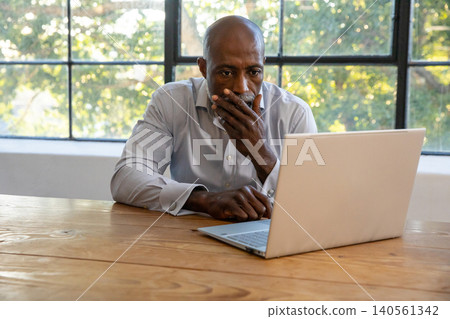 African american man wearing grey shirt sitting at wooden table working on laptop in office African american man wearing grey shirt sitting at wooden table working on laptop in office 140561342
