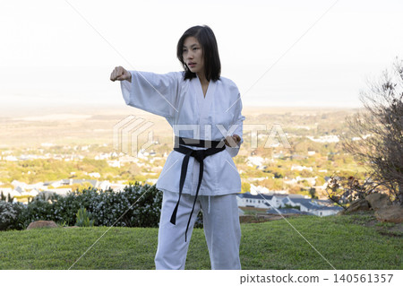Asian woman practicing straight punch stance on grassy hillside wearing white gi and 48belt Asian woman practicing straight punch stance on grassy hillside wearing white gi and 48belt 140561357