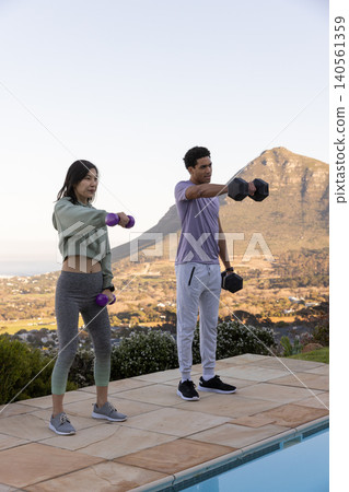 Diverse friends lifting dumbbells on stone patio near swimming pool under clear sky Diverse friends lifting dumbbells on stone patio near swimming pool under clear sky 140561359