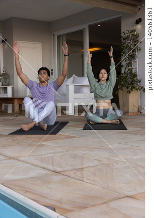 Diverse couple practicing yoga sitting cross-legged raising arms overhead at poolside on mats Diverse couple practicing yoga sitting cross-legged raising arms overhead at poolside on mats 140561361