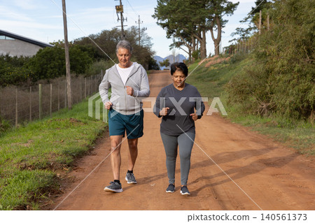 Senior indian couple jogging together on dirt road wearing running shoes, passing wire fence Senior indian couple jogging together on dirt road wearing running shoes, passing wire fence 140561373