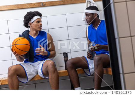 African american male teammates sitting on bench in locker room with basketball, water bottle African american male teammates sitting on bench in locker room with basketball, water bottle 140561375