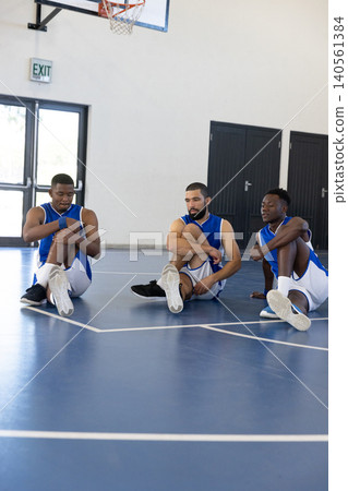 Diverse male teammates in blue uniforms stretching under basketball hoop, exit sign at gym Diverse male teammates in blue uniforms stretching under basketball hoop, exit sign at gym 140561384