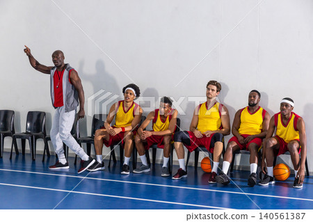 African american coach pointing and five basketball players sitting near basketballs on court African american coach pointing and five basketball players sitting near basketballs on court 140561387
