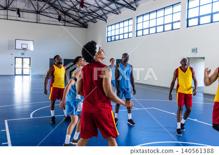 Diverse male basketball players reaching for rebound on blue court in gymnasium under hoop Diverse male basketball players reaching for rebound on blue court in gymnasium under hoop 140561388