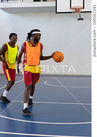 African american teammates in vests dribbling basketball and running on gym court with hoop African american teammates in vests dribbling basketball and running on gym court with hoop 140561391