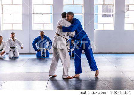 Diverse martial arts students grappling in gis on dojo mats, classmates sitting in seiza 140561395