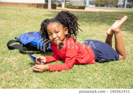 African american child in red top lying on park lawn holding smartphone with backpack African american child in red top lying on park lawn holding smartphone with backpack 140561431