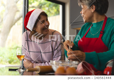 Diverse senior female friends in Santa hat laughing and whisking batter in bowl at kitchen counter Diverse senior female friends in Santa hat laughing and whisking batter in bowl at kitchen counter 140561444