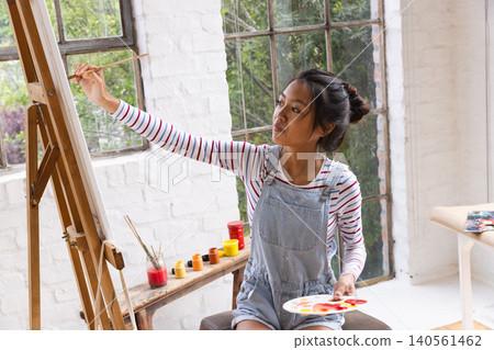 Asian female artist in overalls applying paint to canvas with brush in art studio holding palette Asian female artist in overalls applying paint to canvas with brush in art studio holding palette 140561462