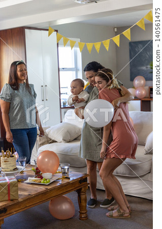 Diverse female friends holding baby boy and hugging in living room with cake, gift, balloons Diverse female friends holding baby boy and hugging in living room with cake, gift, balloons 140561475