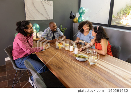 Diverse friends sitting around wooden dining table holding glasses by lit candles, snack bowls Diverse friends sitting around wooden dining table holding glasses by lit candles, snack bowls 140561480