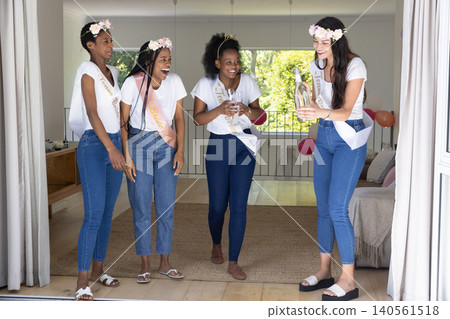 Diverse female friends wearing floral headbands holding champagne flutes near balloons at home Diverse female friends wearing floral headbands holding champagne flutes near balloons at home 140561518