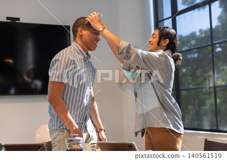 Diverse coworkers lifting coil and placing coil on coworker head near laptops in office Diverse coworkers lifting coil and placing coil on coworker head near laptops in office 140561519