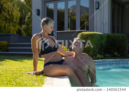 Senior couple sitting on tiled pool edge in backyard wearing black bikini, swim trunks Senior couple sitting on tiled pool edge in backyard wearing black bikini, swim trunks 140561544