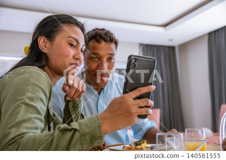 Diverse couple leaning at breakfast table in dining nook with smartphone above glasses of juice Diverse couple leaning at breakfast table in dining nook with smartphone above glasses of juice 140561555