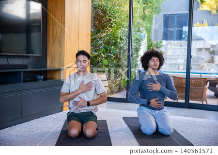 Diverse male friends practicing deep breathing on yoga mats in living room by sliding glass doors Diverse male friends practicing deep breathing on yoga mats in living room by sliding glass doors 140561571