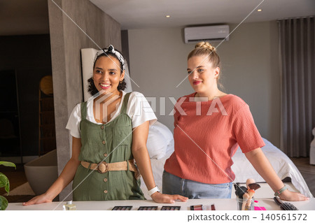 Diverse female friends standing behind countertop in bedroom showcasing makeup palettes, brushes Diverse female friends standing behind countertop in bedroom showcasing makeup palettes, brushes 140561572