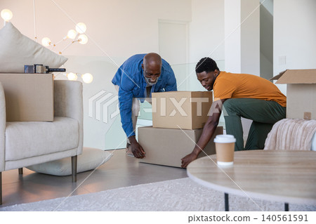 African american father and son lifting cardboard boxes in living room with couch, lamp, coffee cup African american father and son lifting cardboard boxes in living room with couch, lamp, coffee cup 140561591