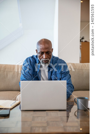 African american senior man working on laptop at coffee table in living room with notebook African american senior man working on laptop at coffee table in living room with notebook 140561593