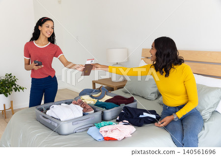 Diverse mom and teenage daughter at home, mother handing passport and ticket into open suitcase Diverse mom and teenage daughter at home, mother handing passport and ticket into open suitcase 140561696