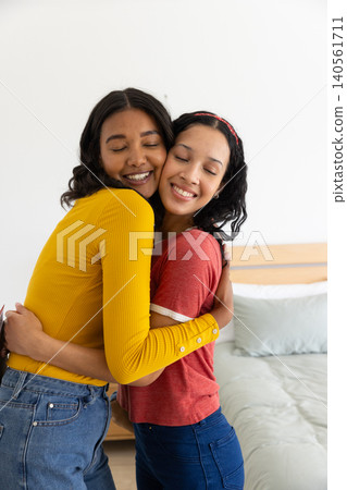 Diverse mom and teenage daughter hugging each other in home bedroom with wooden headboard Diverse mom and teenage daughter hugging each other in home bedroom with wooden headboard 140561711