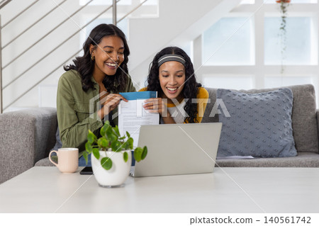 Diverse mom and teenage daughter sitting on sofa in living room holding paper using laptop Diverse mom and teenage daughter sitting on sofa in living room holding paper using laptop 140561742