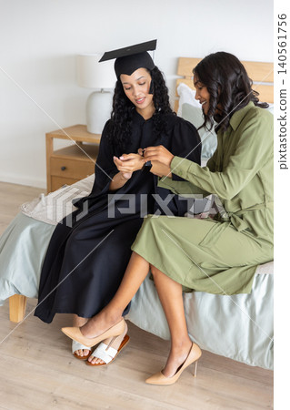 Diverse mom and teenage daughter examining graduation ring on finger, wearing gown and cap Diverse mom and teenage daughter examining graduation ring on finger, wearing gown and cap 140561756